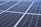 A close-up view of a large array of solar panels, with a uniform pattern of dark blue photovoltaic cells arranged in a grid. The panels are lined with silver frames, reflecting sunlight and indicating a clear day.
