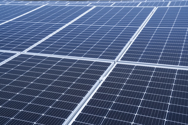 A close-up view of a large array of solar panels, with a uniform pattern of dark blue photovoltaic cells arranged in a grid. The panels are lined with silver frames, reflecting sunlight and indicating a clear day.