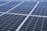 A close-up view of a large array of solar panels, with a uniform pattern of dark blue photovoltaic cells arranged in a grid. The panels are lined with silver frames, reflecting sunlight and indicating a clear day.
