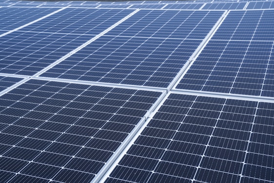 A close-up view of a large array of solar panels, with a uniform pattern of dark blue photovoltaic cells arranged in a grid. The panels are lined with silver frames, reflecting sunlight and indicating a clear day.