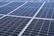 A close-up view of a large array of solar panels, with a uniform pattern of dark blue photovoltaic cells arranged in a grid. The panels are lined with silver frames, reflecting sunlight and indicating a clear day.