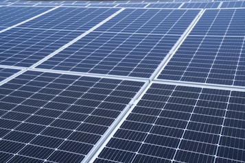 A close-up view of a large array of solar panels, with a uniform pattern of dark blue photovoltaic cells arranged in a grid. The panels are lined with silver frames, reflecting sunlight and indicating a clear day.