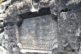 An aged stone plaque with Latin inscriptions is embedded in a rough, weathered stone wall. The text appears worn, indicating the historical nature of the monument.