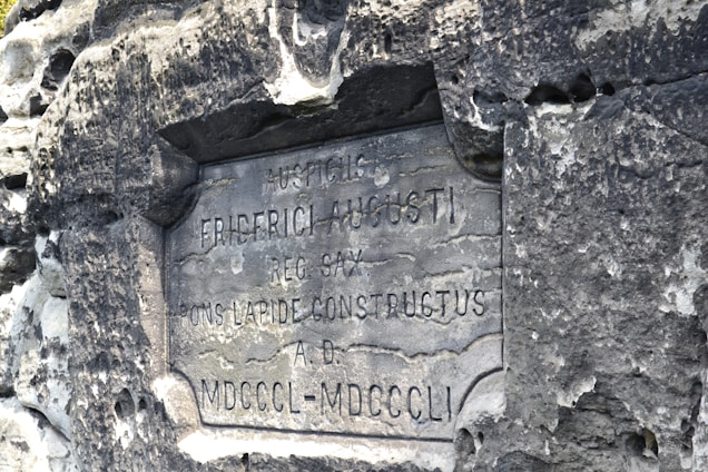 An aged stone plaque with Latin inscriptions is embedded in a rough, weathered stone wall. The text appears worn, indicating the historical nature of the monument.