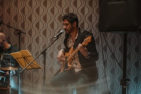 A musician plays an electric guitar while sitting on a stool in a dimly lit space with a patterned wallpaper in the background. A microphone is positioned in front of him, suggesting live music performance. Another individual, possibly a band member, is visible in the background near a music stand with yellow sheet music.