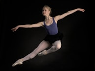 A ballet dancer captured in mid-air performing a graceful leap. The dancer is wearing a blue leotard, white tights, and pointe shoes. Her arms are extended elegantly, and her expression conveys focus and poise. The background is dark, highlighting the dancer's form.