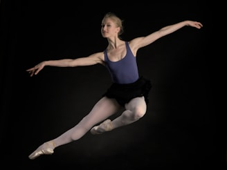 A ballet dancer captured in mid-air performing a graceful leap. The dancer is wearing a blue leotard, white tights, and pointe shoes. Her arms are extended elegantly, and her expression conveys focus and poise. The background is dark, highlighting the dancer's form.