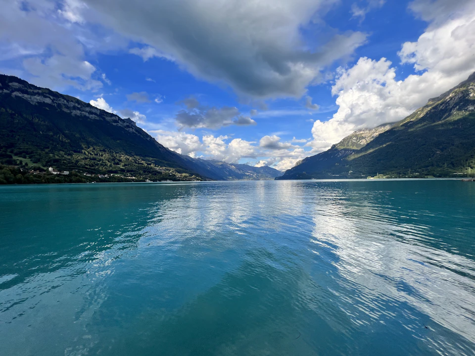 The image depicts a serene lake surrounded by majestic mountains, with a clear blue sky above and fluffy white clouds below. The calm water reflects the surrounding landscape, creating a mirror-like effect that enhances the beauty of the scene. The perspective is from the shoreline, looking out towards the mountains in the distance, providing a sense of depth and vastness to the image.