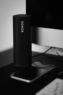A sleek, cylindrical black speaker with the brand name SONOS in white letters stands upright next to a large screen monitor. Below it, a smartphone with a dark screen rests on the surface.