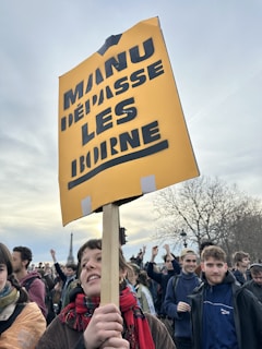A group of people participating in a protest or demonstration. A person in the foreground is holding a yellow sign with the words 'MANU DEpASSE LES Borne' written in bold, black letters. There is a crowd of individuals in the background, some with raised hands, indicating an active and engaging event. The atmosphere appears energetic and peaceful.