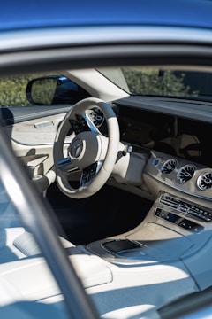 The interior of a modern car featuring a sophisticated design with a beige and black color scheme. The steering wheel has a logo at the center, and the dashboard includes multiple air vents, a digital display, and various control buttons. The driver’s seat and part of the passenger seat are visible, both upholstered in high-quality material. Sunlight casts onto the seats and the center console, creating a bright and airy atmosphere.