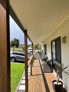 A wooden porch extends along the side of a house with white exterior walls. The porch has a wooden railing and steps leading down to a well-maintained grassy area. Outdoor furniture, including chairs and a potted plant, are neatly arranged on the porch. A car is parked on the driveway adjacent to the porch. Trees and additional greenery are visible in the background under a partly cloudy sky.