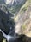 The photograph captures a stunning landscape featuring a deep valley with a flowing river cutting through rugged cliffs. There is a waterfall visible in the lower portion of the image, with mist rising and creating a partial rainbow. The valley is lush with green vegetation, and the rocky cliffs are covered in patches of greenery.