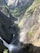 The photograph captures a stunning landscape featuring a deep valley with a flowing river cutting through rugged cliffs. There is a waterfall visible in the lower portion of the image, with mist rising and creating a partial rainbow. The valley is lush with green vegetation, and the rocky cliffs are covered in patches of greenery.