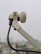 A close-up of a white satellite dish arm and its attached components, including a cable that stretches downward. The background shows an urban landscape with buildings that appear slightly blurred due to depth of field.
