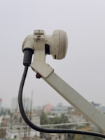 A close-up of a white satellite dish arm and its attached components, including a cable that stretches downward. The background shows an urban landscape with buildings that appear slightly blurred due to depth of field.