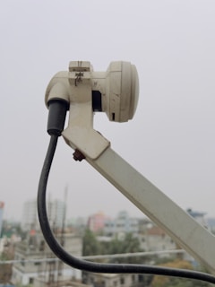 A close-up of a white satellite dish arm and its attached components, including a cable that stretches downward. The background shows an urban landscape with buildings that appear slightly blurred due to depth of field.