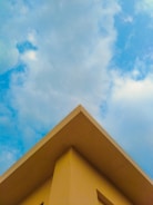 A triangular rooftop against a backdrop of a partly cloudy sky. The roof is painted in a soft yellow color, creating a contrast with the blue sky and white clouds.