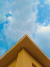 A triangular rooftop against a backdrop of a partly cloudy sky. The roof is painted in a soft yellow color, creating a contrast with the blue sky and white clouds.