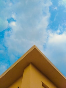 A triangular rooftop against a backdrop of a partly cloudy sky. The roof is painted in a soft yellow color, creating a contrast with the blue sky and white clouds.
