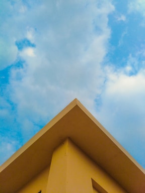 A triangular rooftop against a backdrop of a partly cloudy sky. The roof is painted in a soft yellow color, creating a contrast with the blue sky and white clouds.