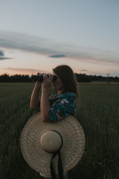 A person stands in a field during twilight, holding binoculars and looking into the distance. They are wearing a large straw hat slung over their back and a colorful, patterned shirt. The sky is clear with soft clouds and the sun is setting on the horizon, casting a muted glow across the landscape.