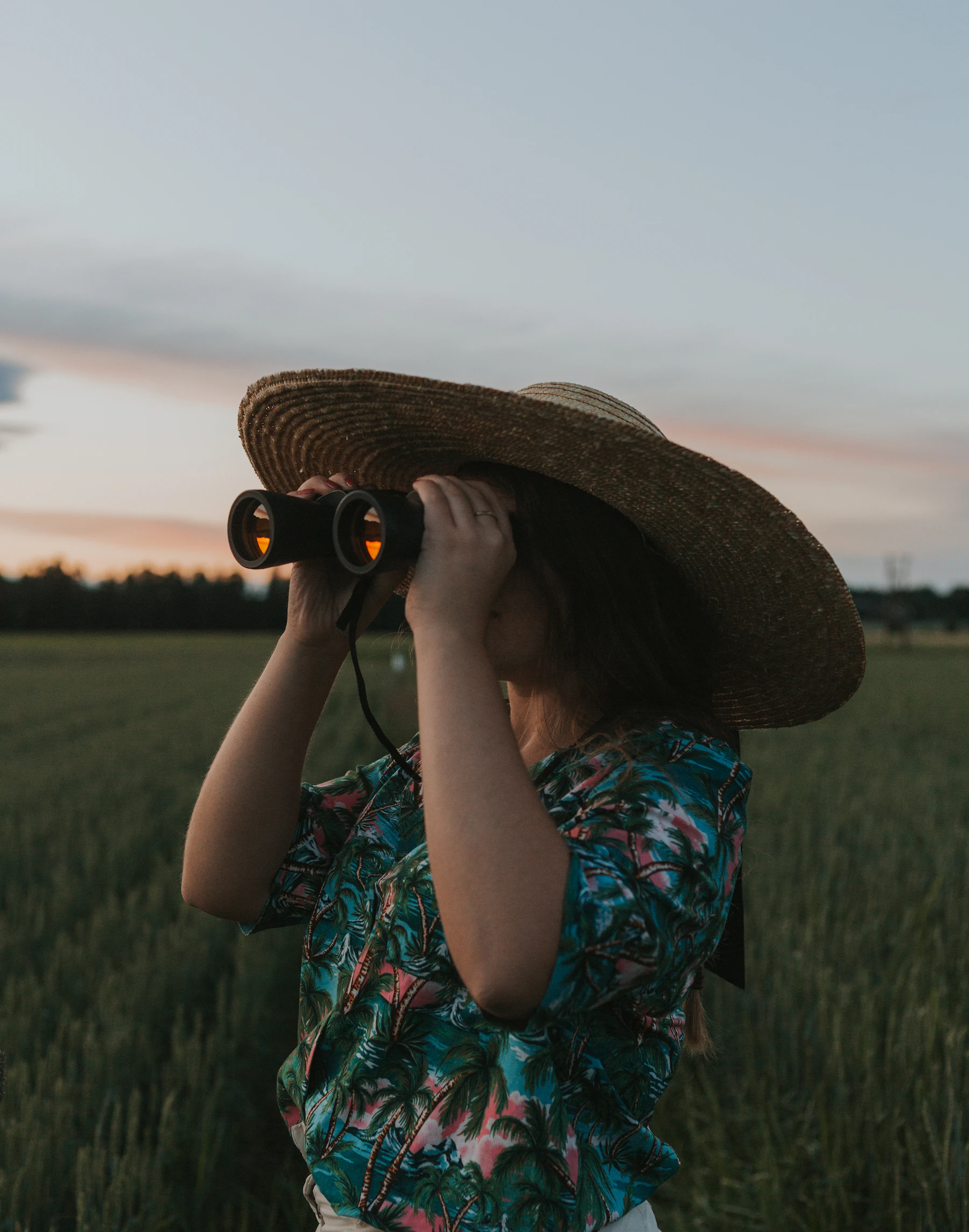 A person in a wide straw hat looking through binoculars across a green field