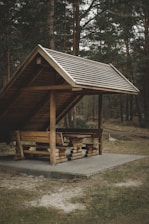 A wooden shelter with a slanted roof stands in a wooded area. The structure is made of logs and includes a picnic table and benches underneath the roof. Surrounding the shelter are tall trees, creating a secluded and natural environment.