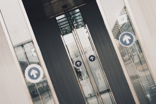 Several glass doors, likely part of a building entrance or exit, display circular blue signs with upward-facing arrows. The doors are slightly open, revealing a partial view through them to the other side. There are reflections on the glass panels of the surrounding environment, including a building and some pavement. The scene has an industrial and urban feel.