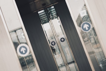 Several glass doors, likely part of a building entrance or exit, display circular blue signs with upward-facing arrows. The doors are slightly open, revealing a partial view through them to the other side. There are reflections on the glass panels of the surrounding environment, including a building and some pavement. The scene has an industrial and urban feel.