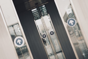 Several glass doors, likely part of a building entrance or exit, display circular blue signs with upward-facing arrows. The doors are slightly open, revealing a partial view through them to the other side. There are reflections on the glass panels of the surrounding environment, including a building and some pavement. The scene has an industrial and urban feel.