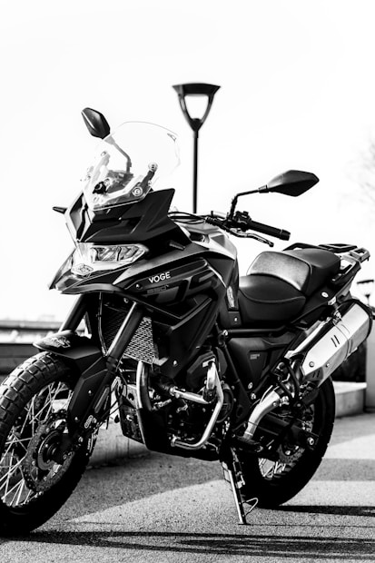 A sleek motorcycle stands on a paved surface, showcasing its detailed design. The bike features rugged tires and a clear windshield, complemented by a sturdy frame and side mirrors. In the background, a lamp post is visible against an overcast sky.