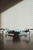 A small, light gray drone with four rotors is positioned on a dark wooden surface. The shadows of window blinds create diagonal lines across the background wall, adding a pattern to the minimalist scene.