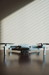 A small, light gray drone with four rotors is positioned on a dark wooden surface. The shadows of window blinds create diagonal lines across the background wall, adding a pattern to the minimalist scene.