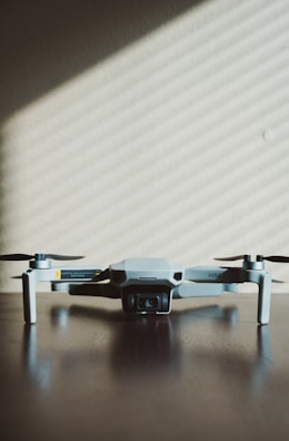 A small, light gray drone with four rotors is positioned on a dark wooden surface. The shadows of window blinds create diagonal lines across the background wall, adding a pattern to the minimalist scene.