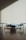 A small, light gray drone with four rotors is positioned on a dark wooden surface. The shadows of window blinds create diagonal lines across the background wall, adding a pattern to the minimalist scene.
