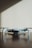 A small, light gray drone with four rotors is positioned on a dark wooden surface. The shadows of window blinds create diagonal lines across the background wall, adding a pattern to the minimalist scene.