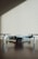 A small, light gray drone with four rotors is positioned on a dark wooden surface. The shadows of window blinds create diagonal lines across the background wall, adding a pattern to the minimalist scene.