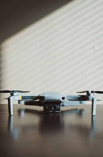 A small, light gray drone with four rotors is positioned on a dark wooden surface. The shadows of window blinds create diagonal lines across the background wall, adding a pattern to the minimalist scene.