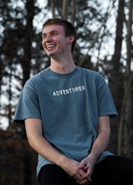 A young man is smiling broadly while sitting outdoors. He is wearing a blue t-shirt with the word 'Adventurer' printed on it. The background is filled with trees, creating a serene natural setting.