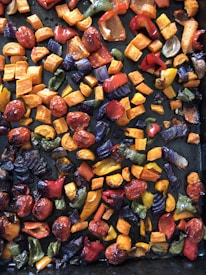 A colorful assortment of roasted vegetables including chunks of red, yellow, and green peppers, along with cherry tomatoes and diced purple onions scattered on a dark baking tray.