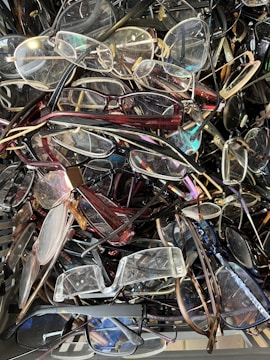 A large pile of various eyeglasses, including different styles and colors of frames and lenses. The collection appears random and disorganized, with some glasses overlapped and tangled.