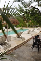 A serene outdoor pool area featuring several white lounge chairs lined up on a clean deck with lush green tropical plants surrounding the space. The pool itself is a soft blue color, creating a tranquil atmosphere. Palm fronds and other foliage partially frame the view, adding a sense of seclusion to the setting. A wooden table with black metal chairs is seen nearby on a concrete surface, suggesting a spot for relaxation or dining.