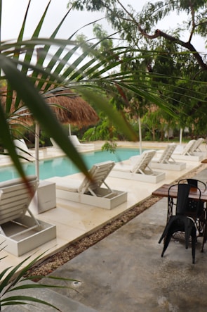 A serene outdoor pool area featuring several white lounge chairs lined up on a clean deck with lush green tropical plants surrounding the space. The pool itself is a soft blue color, creating a tranquil atmosphere. Palm fronds and other foliage partially frame the view, adding a sense of seclusion to the setting. A wooden table with black metal chairs is seen nearby on a concrete surface, suggesting a spot for relaxation or dining.
