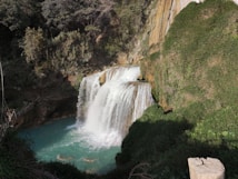 A picturesque waterfall cascades into a turquoise pool surrounded by lush green vegetation and rocky cliffs. The water flows with vigor, creating mist at the base. The surrounding area is dense with trees and foliage, giving a sense of untouched nature.