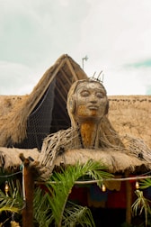 A large woven sculpture of a human head and upper body is positioned on top of a thatched roof structure. The sculpture is made from natural materials like wood and straw, giving it an organic appearance. Surrounding the structure are green palm plants, and the sky in the background is lightly clouded. Strings of lights add a decorative touch to the scene.