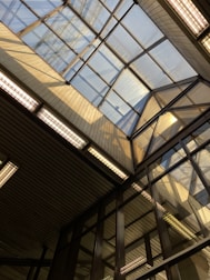 A structural view featuring a large glass ceiling with a geometric design. Sunlight streams through the glass, casting shadows and illuminating the surrounding area. The construction consists of metal and glass, creating a modern architectural aesthetic.