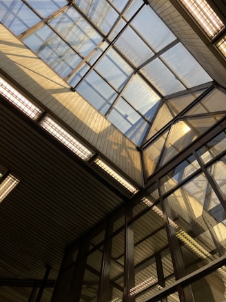 A structural view featuring a large glass ceiling with a geometric design. Sunlight streams through the glass, casting shadows and illuminating the surrounding area. The construction consists of metal and glass, creating a modern architectural aesthetic.