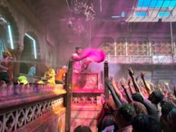 A vibrant celebration scene with a group of people gathered in a richly decorated setting. One person, elevated on a podium, is throwing pink colored powder into the air, while the crowd below reaches up towards it. There are decorations and lights, and the architecture is ornate with intricate designs and a chandelier hanging from the ceiling.