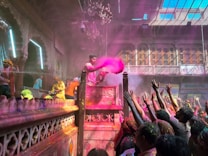 A vibrant celebration scene with a group of people gathered in a richly decorated setting. One person, elevated on a podium, is throwing pink colored powder into the air, while the crowd below reaches up towards it. There are decorations and lights, and the architecture is ornate with intricate designs and a chandelier hanging from the ceiling.